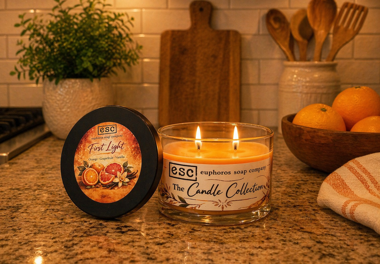 Orange candle with packaging on a kitchen counter, featuring oranges and utensils in the background. The candle has two wicks that are lit.