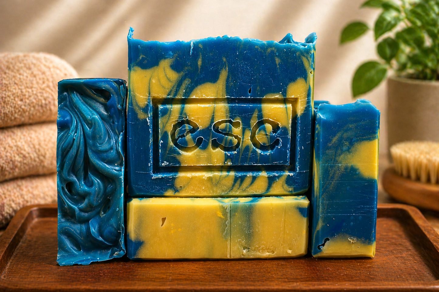 Four handcrafted yellow and blue marbled soap bars arranged on a wooden surface, with the center bar stamped “ESC,” representing Euphoros Soap Company, set against a soft, spa-inspired background.