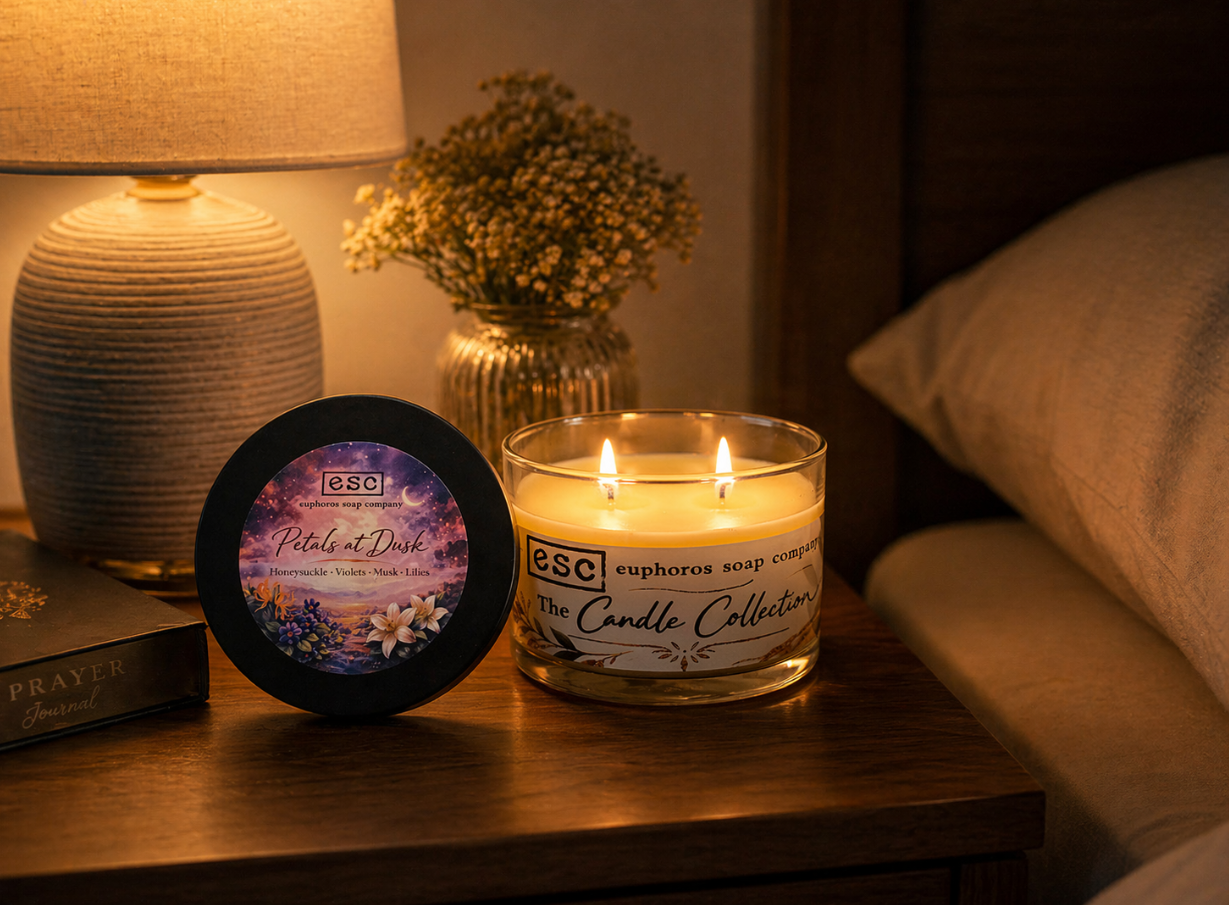 White candle and packaging on a bedside table with a lamp and flowers in the background.  The candle has two wicks that are lit.