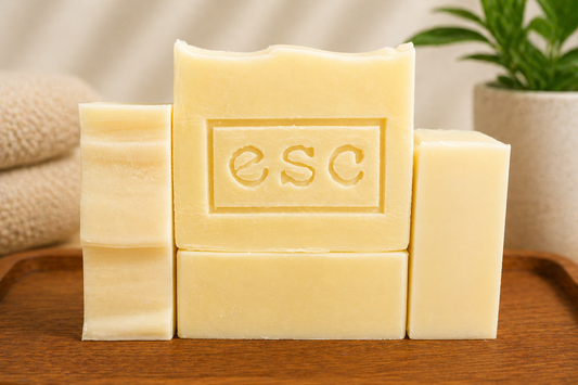 4 bars of white soap with the ESC brand logo imprinted on the center top bar on a wooden surface.  A plant and a towel are in the background. Featuring the brand 'ESC', which is the logo for Euphoros Soap Company.